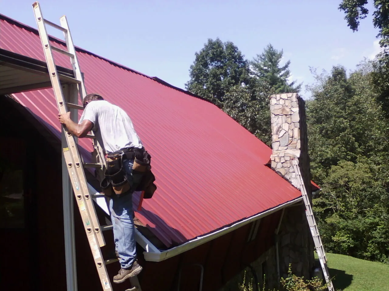 Roofer on a ladder beside a red metal roof and stone chimney — Soffit & Fascia Repair by Roofing Brothers in Waupaca